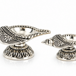 Pair of Mohnaa Jyotika sterling silver ornate diyas displayed in two sizes, featuring decorative elongated bowls and sculpted pedestal bases.