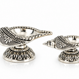 Pair of Mohnaa Jyotika sterling silver ornate diyas displayed in two sizes, featuring decorative elongated bowls and sculpted pedestal bases.