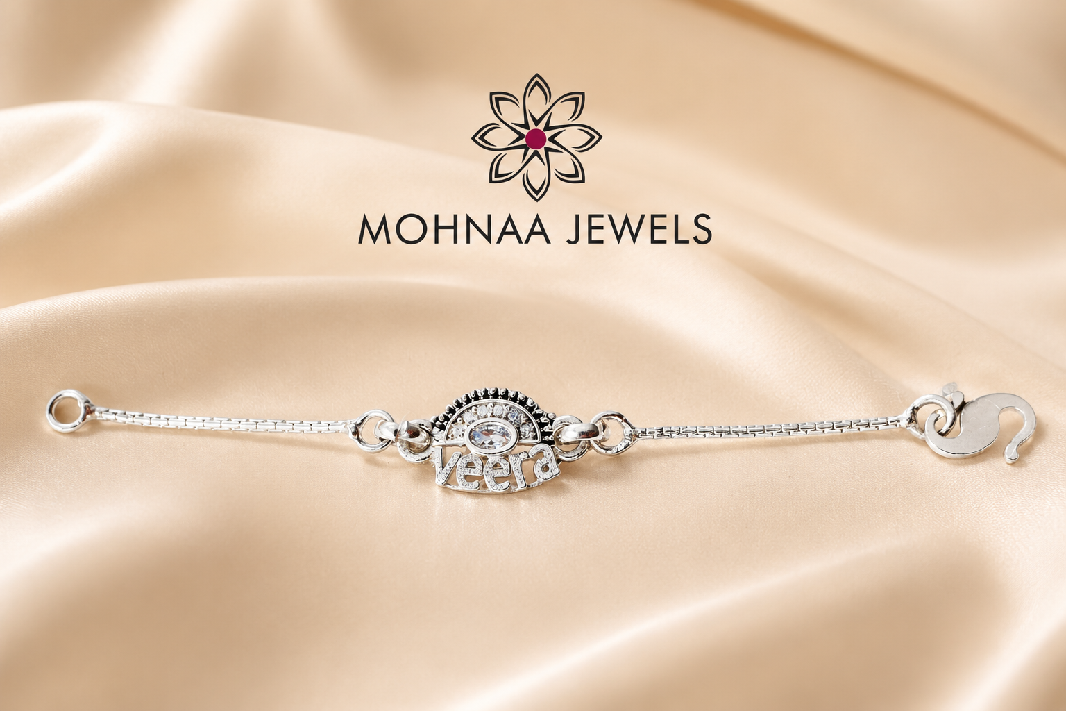 A silver-toned chain bracelet with a central evil eye charm and a small 'VEERA' text charm, displayed on draped champagne-colored satin fabric. The brand logo 'MOHNAO JEWELS' is visible above the bracelet.
